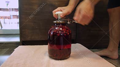 A man canning cherry compote using a manual seaming machine to seal a large glass jar with metal lid. Home canning, food