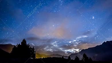 Timelapse night sky stars and moon across fast clouds with mountain background