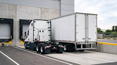 White semi truck trailer parked at loading dock of modern distribution warehouse under cloudy sky