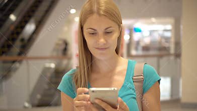 Young woman standing in shopping mall using smartphone, browsing, reading news, chatting with friends