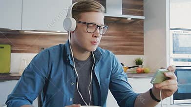 Young laughing man watching streaming movie on smartphone eating cereals with headphones