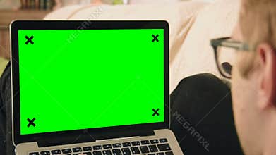 Young man laying on the sofa looking at laptop computer with green screen
