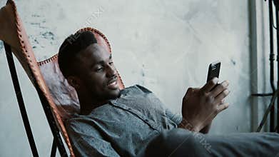 Portrait of African man sitting in chair, using a Smartphone. Handsome male smiles and looks at photos in his phone.
