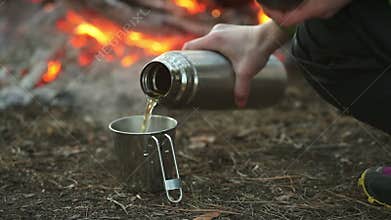 Camper woman camping pours tea from a thermos and drinking in forest near fire