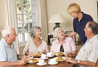 Group Of Senior Couples Enjoying Meal Together In Care Home With Home Help
