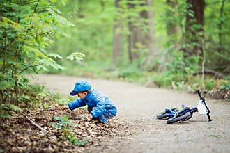 Caucasian little boy toddler in blue jacket, jeans and baseball cap with bike in park playground outside, sitting on ground