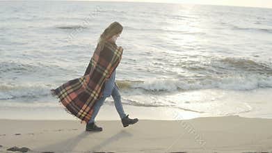 Lonely woman walking on the sandy beach with plaid. Young female spending time on the shore of the sea in cold day.