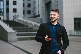 Outdoor portrait of a young businessman who genuinely smiling and holding a smartphone in hand. Business concept.