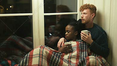 Lovely multi-race couple lying under the red plaid on the window-sill. The man with red hair and beard is drinking tea