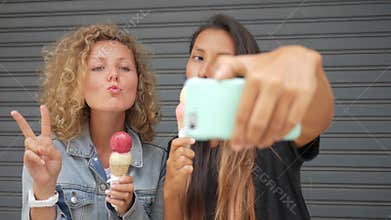 Two young carefree mixed race hipster girls eating ice cream and posing for a selfie photo using mobile phone. lifestyle