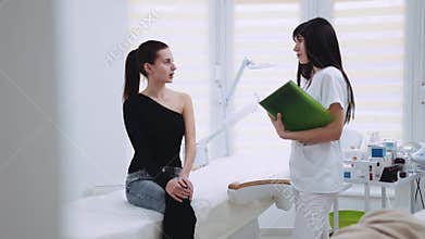 Young brunette woman in elegant wear sits in the cosmetologist’s cabinet, the beautician examines her face, they