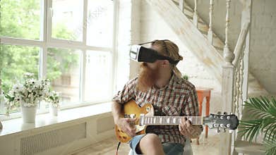 Young bearded man sitting on chair learning to play guitar using VR 360 headset and feels him guitarist at concert at