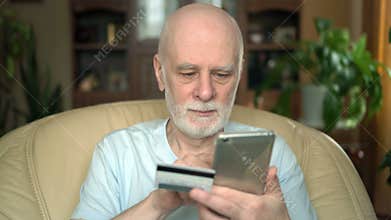 Handsome smiling senior man sitting on chair at home. Buying online with credit card on smartphone