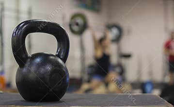 A cast iron kettle bell with a young women weight lifting in the background