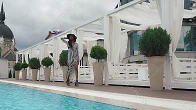 Slow-motion. elegant african american female model in cocktail striped dress and black hat walks at the luxury pool