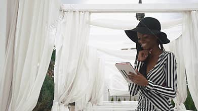 Glamorous african american lady in a striped cocktail dress and black hat using her tablet computer on the luxury hotel