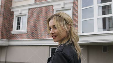 Young woman walking on the street. Blonde turns flirting gaze with the viewer. Black leather jacket
